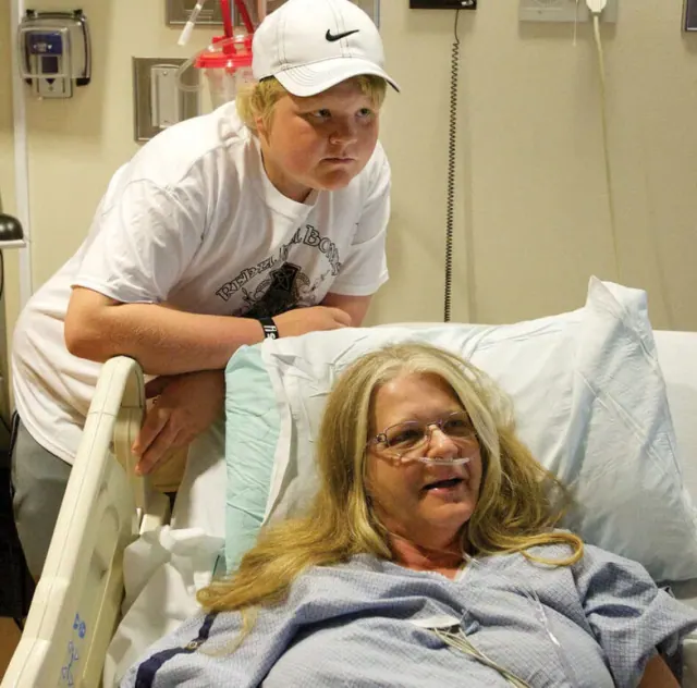 A boy standing beside a woman in a hospital bed