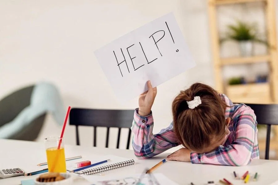 Young girl with her head resting on the table holding a handwritten sign that reads, “HELP!”