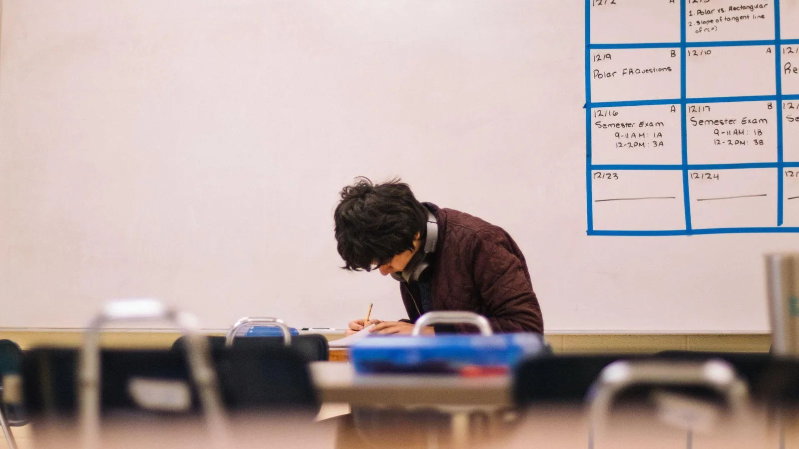 A student with a maroon jacket and headphones sits at a desk in a classroom, intently working on a paper