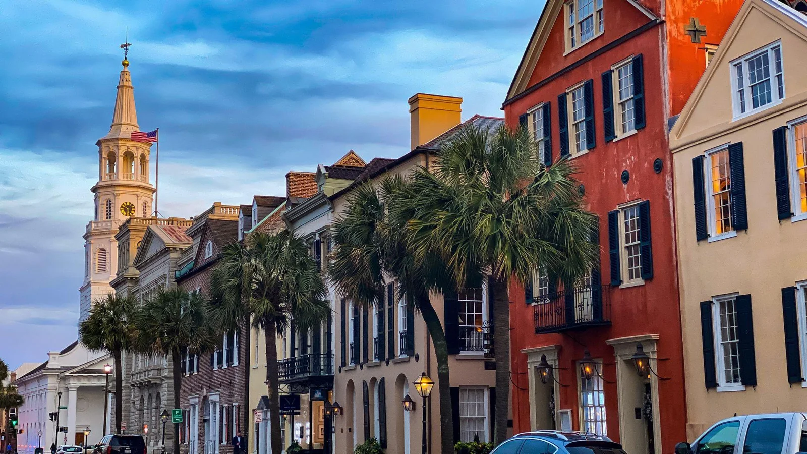 Photograph of a historic street in Charleston, South Carolina, during twilight. The image features a row of traditional Charleston townhouses with colorful facades, black shutters, and palmetto trees in the foreground
