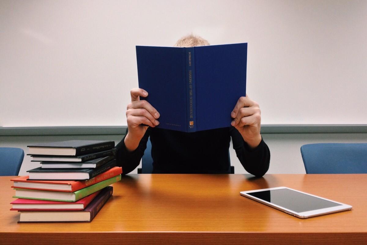 A student holding an open book at a desk beside a stack of books