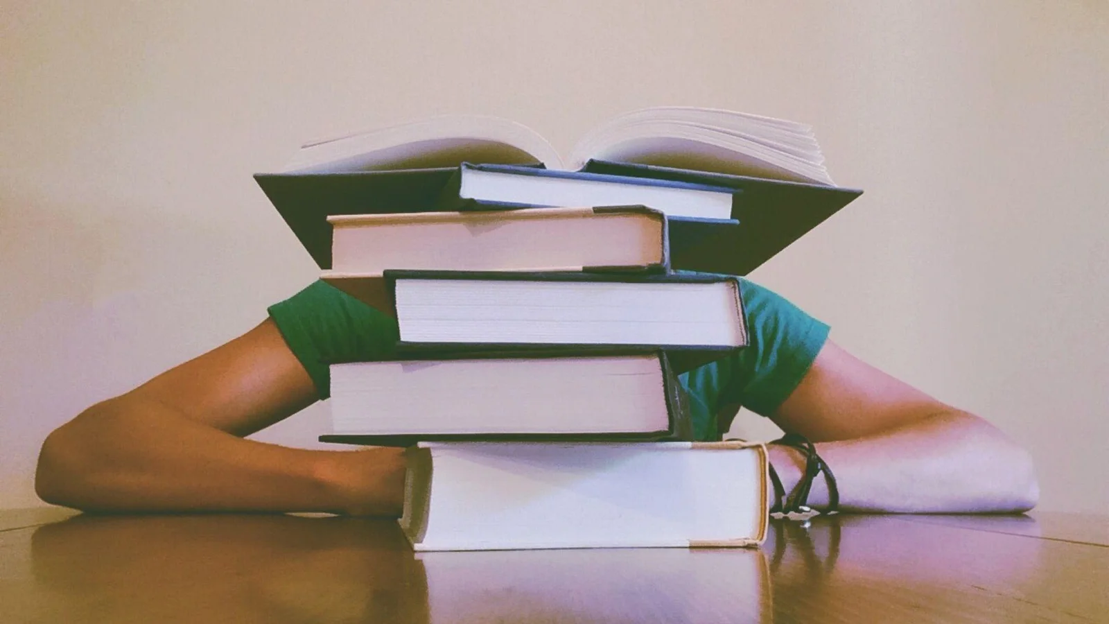A person's arm is extended on a table, being overwhelmed by a stack of books placed on top of the arm