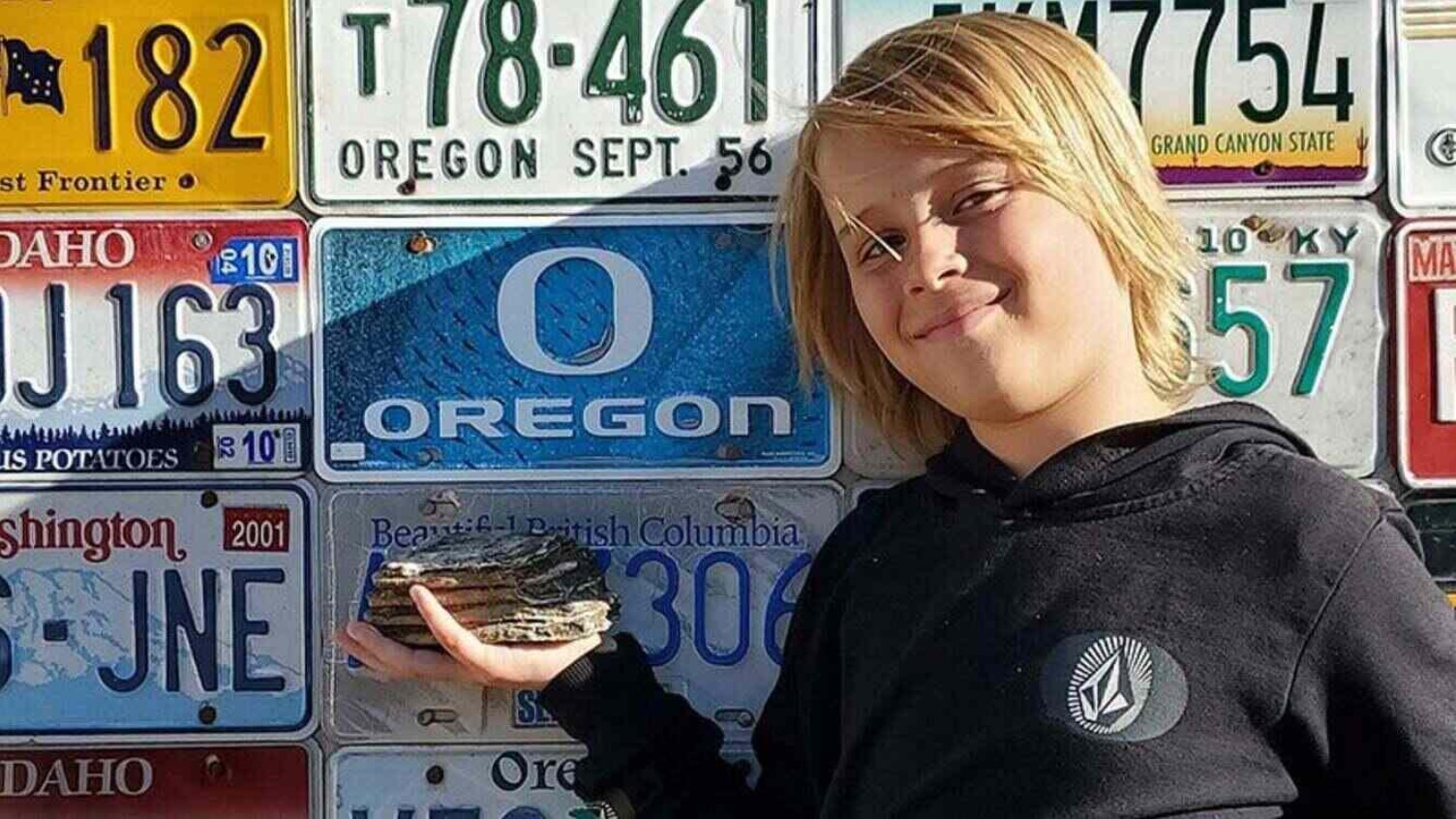 A boy holding an object in front of a wall decorated with vehicle licence plates.