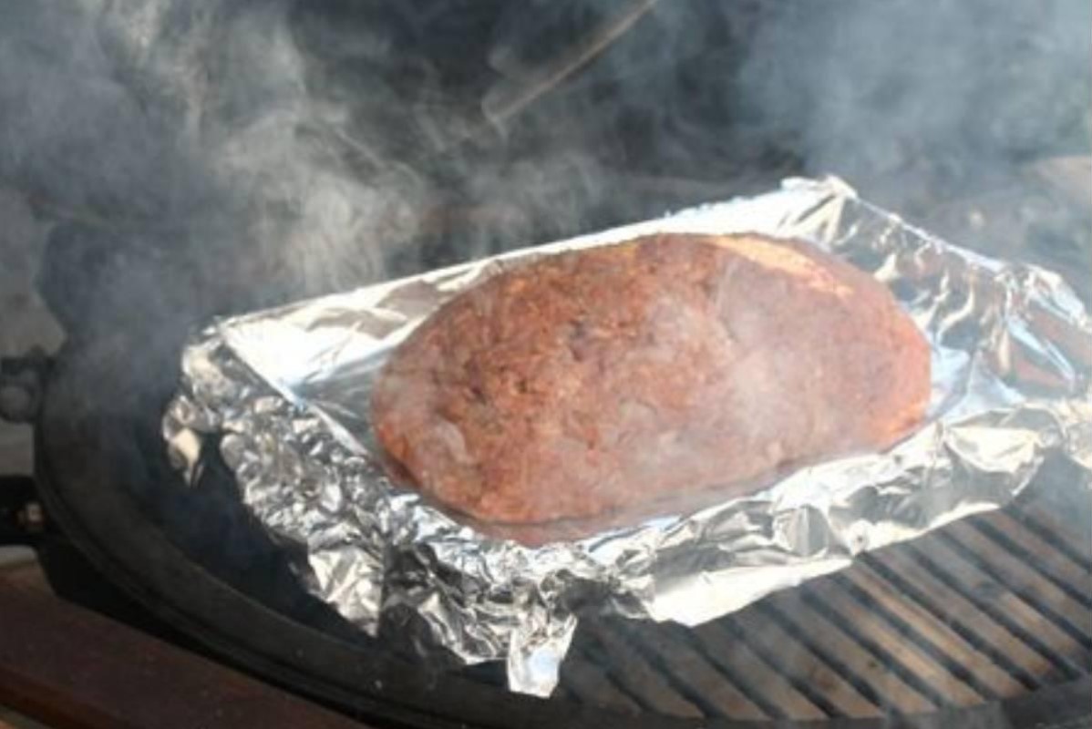 Seasoned meat smoking in a foil-lined tray on a grill.