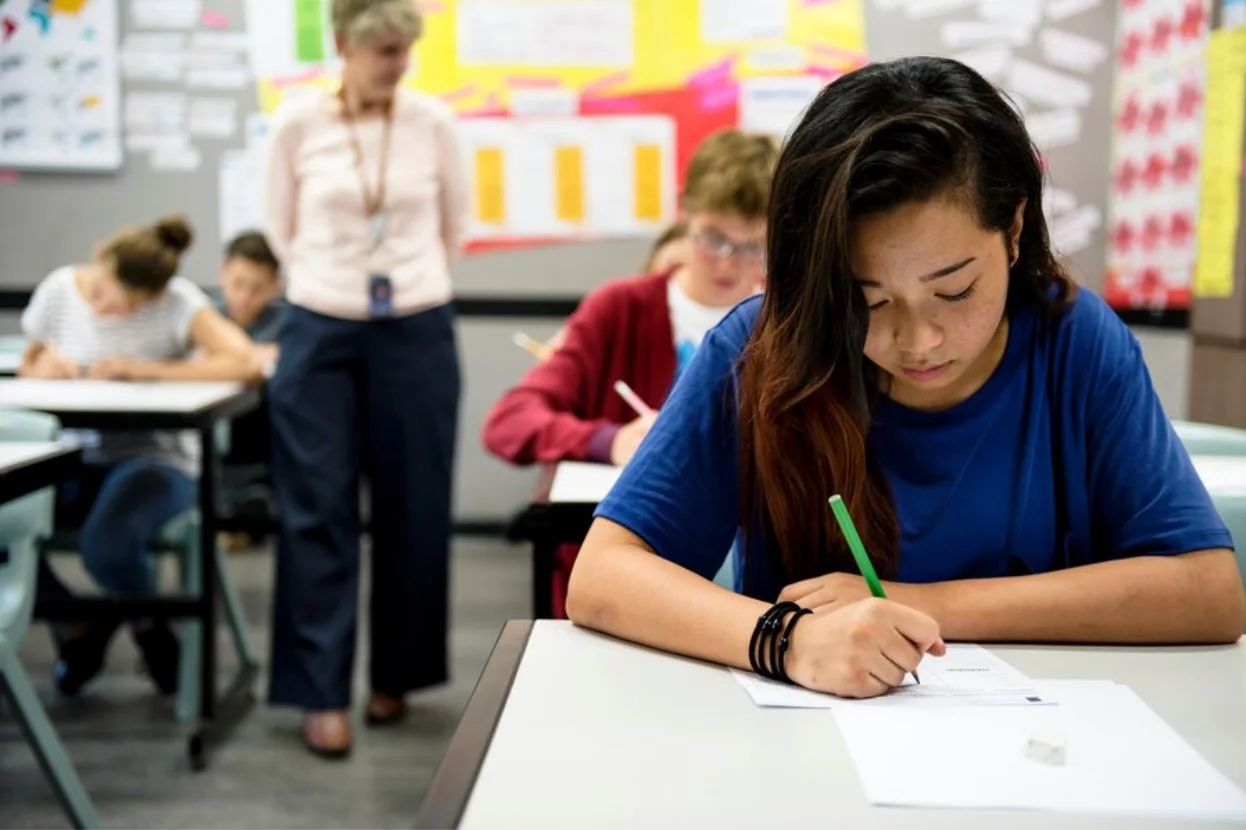 Students taking tests in a classroom while the teacher walks through the aisle