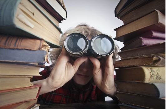 A person looking through binoculars among books, an illustrative photograph