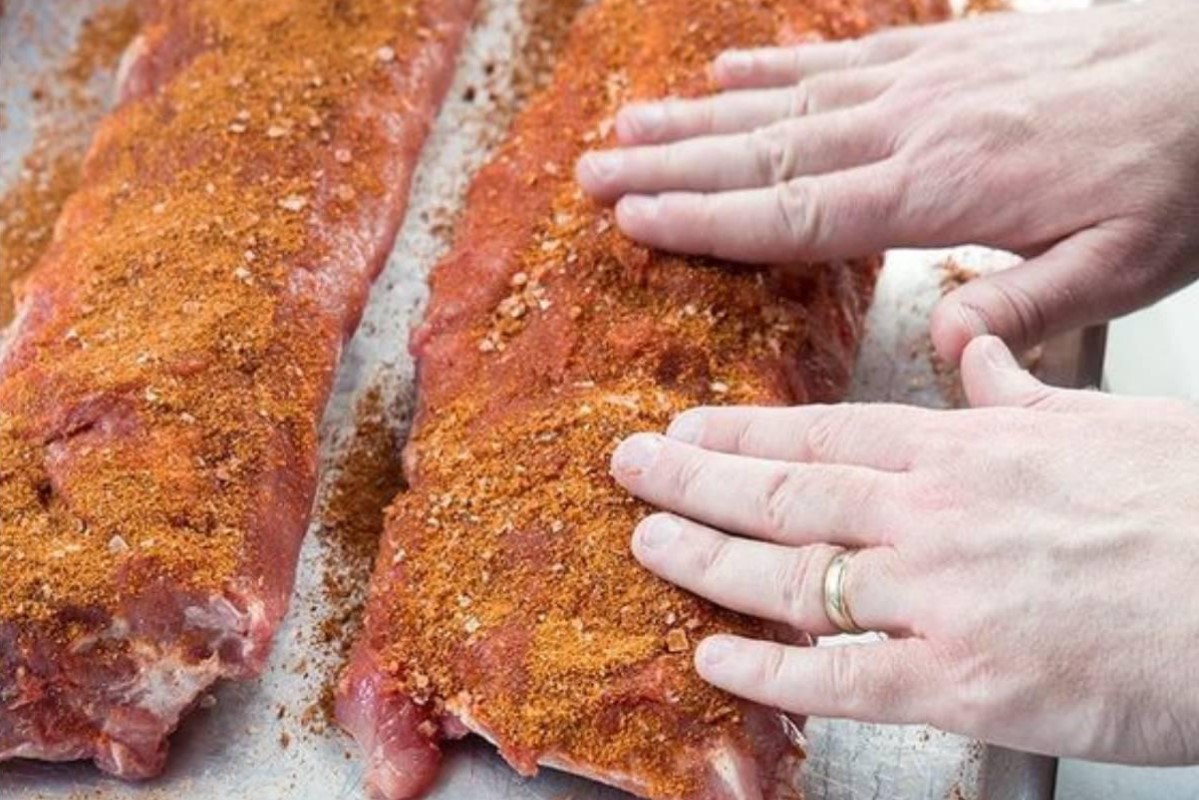 Hands applying a dry seasoning rub to meat.