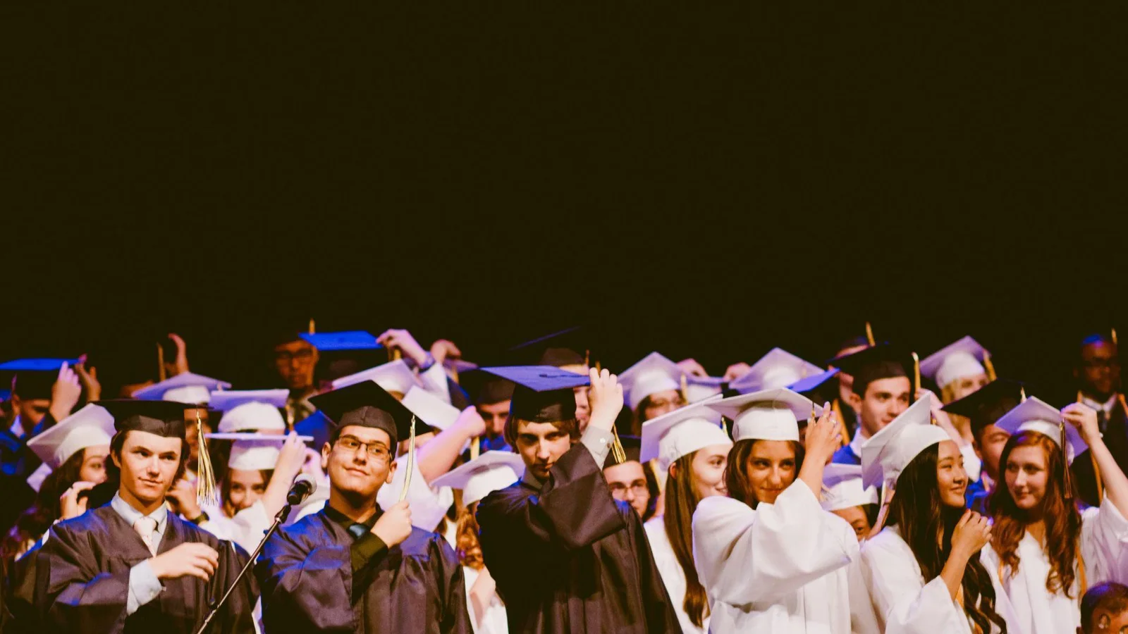 A group of graduates wearing caps and gowns, some in black and some in white, moving their tassels during a graduation ceremony