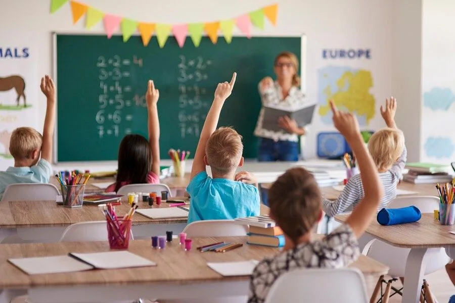 Kids raising their hands in a classroom while the teacher points to one to answer