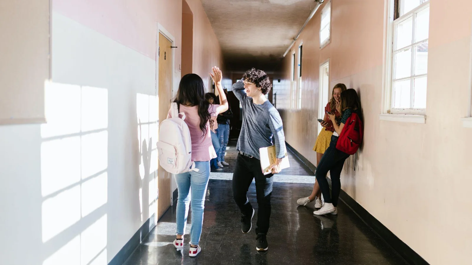 A group of high school students walking and interacting in a school hallway illuminated by natural light from windows