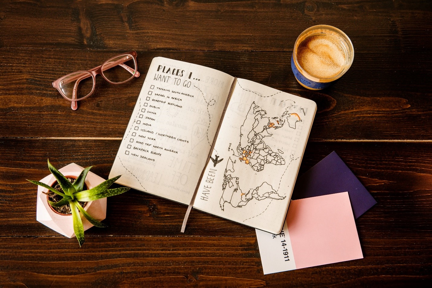 A travel notebook, map, glasses and coffee arranged on a wooden table.