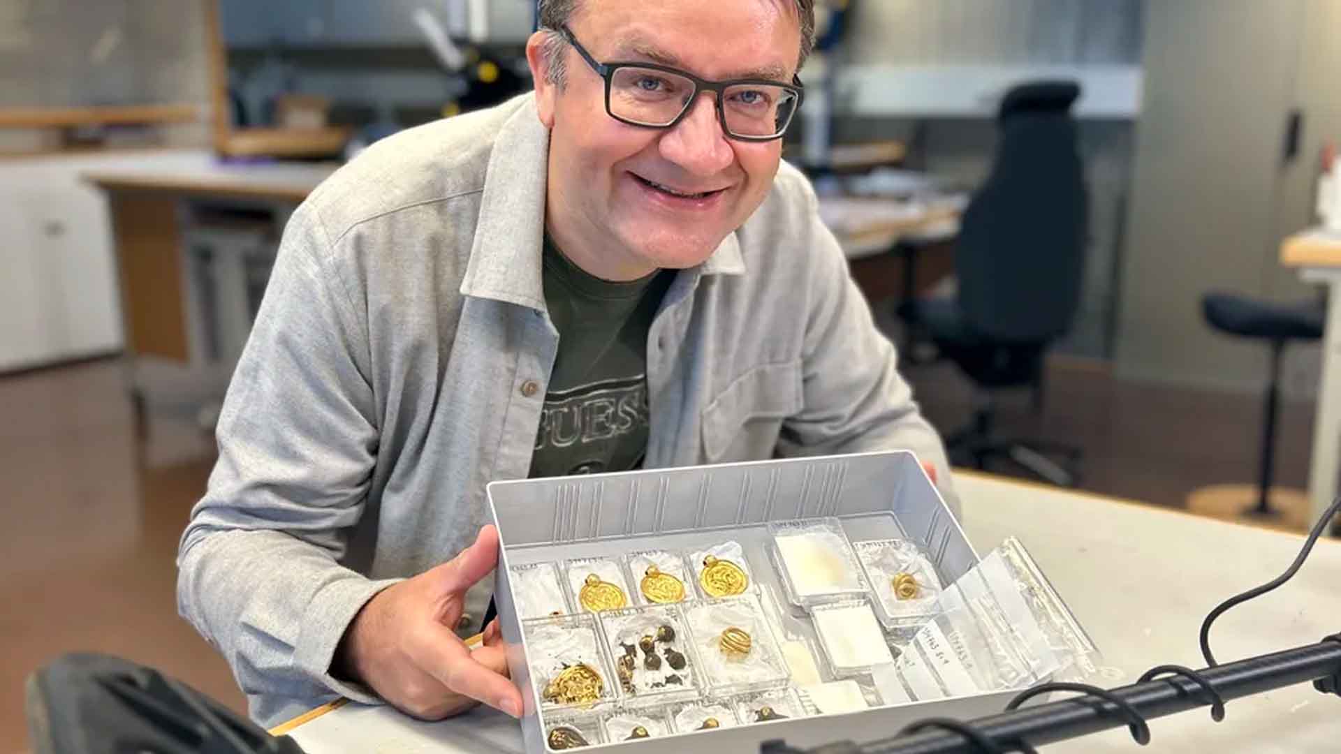 A man holding a display tray of gold objects in an archaeology museum photograph.