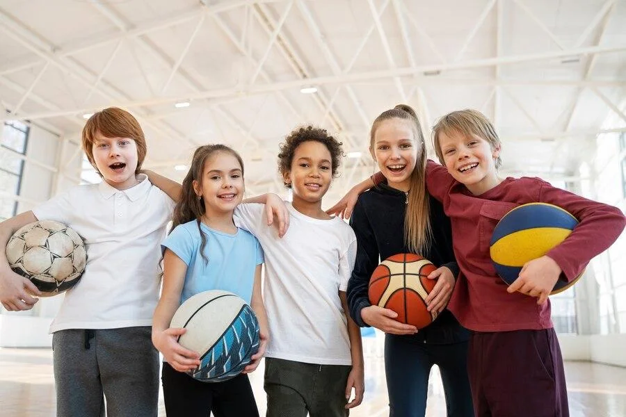 Kids hugging in a gymnasium holding a variety of sports equipment