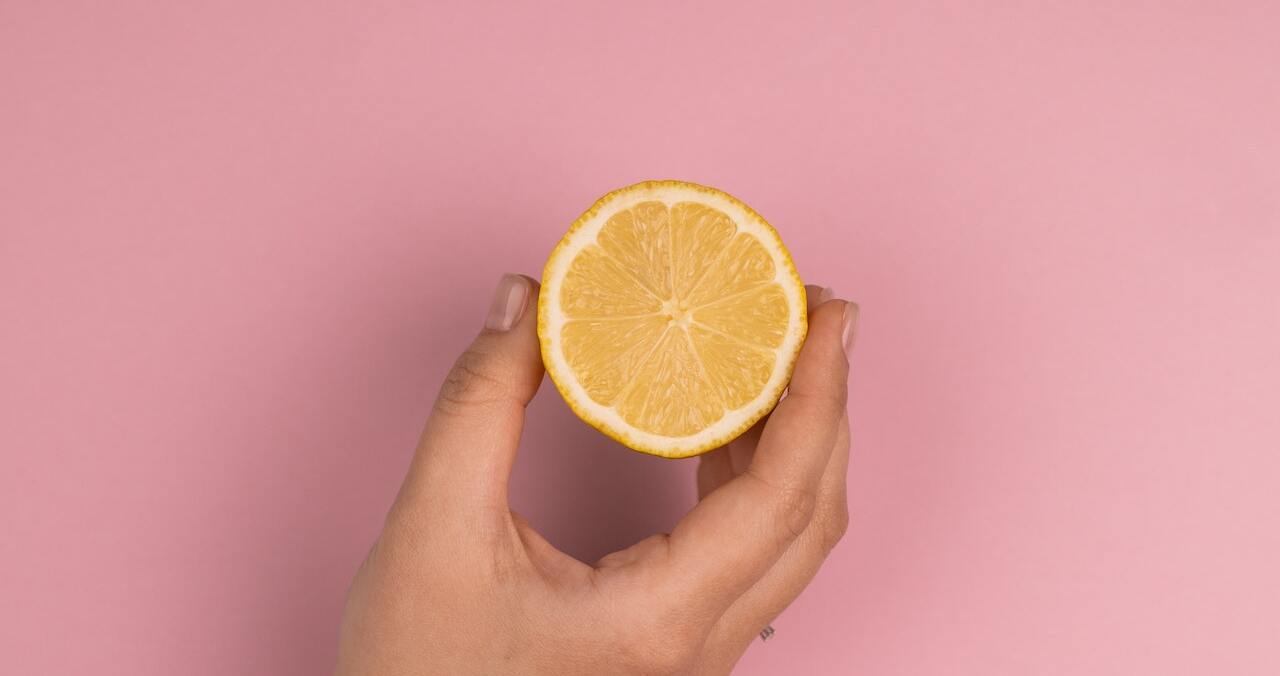 A hand holding a halved lemon against a pink background.