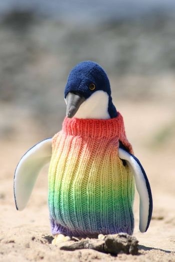 A plush penguin toy in a rainbow-coloured knitted jumper on sand.