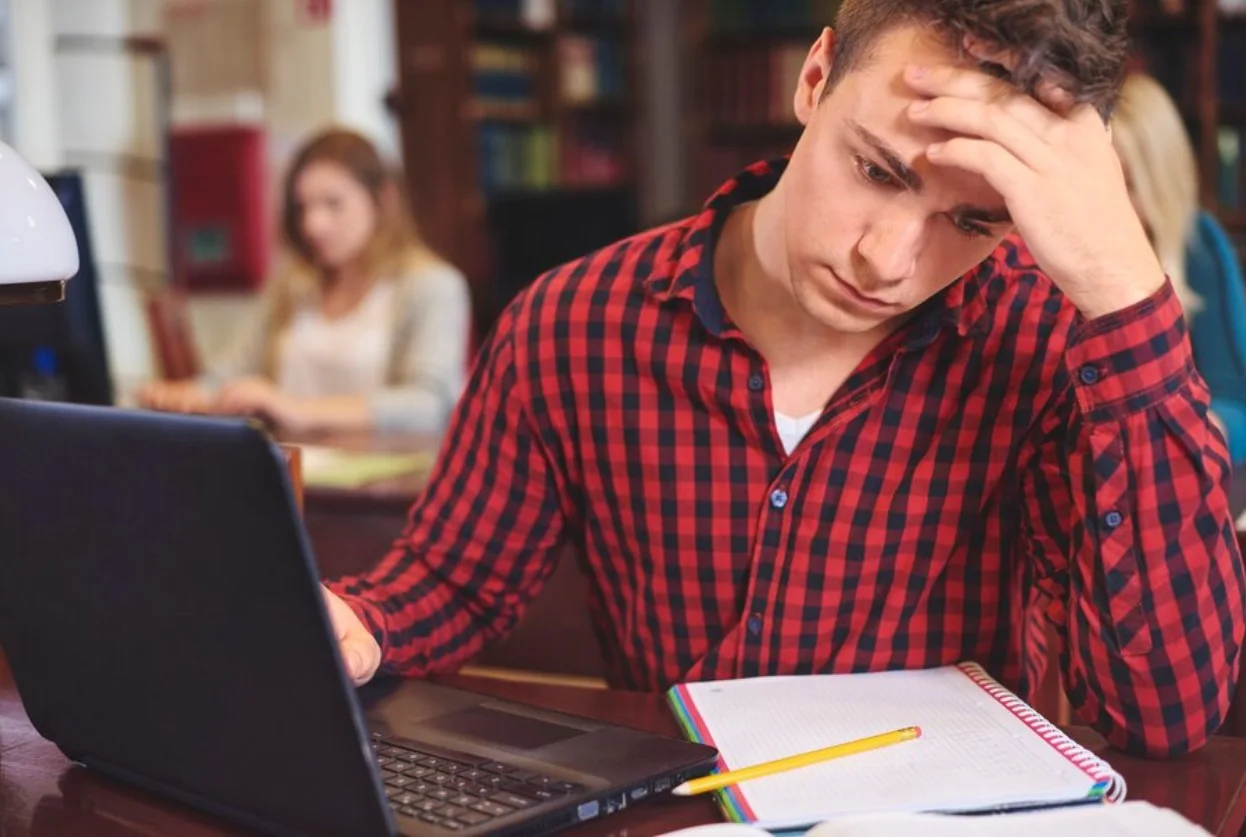 Young man wearing checkered shirt looking down at paper and computer while stressed