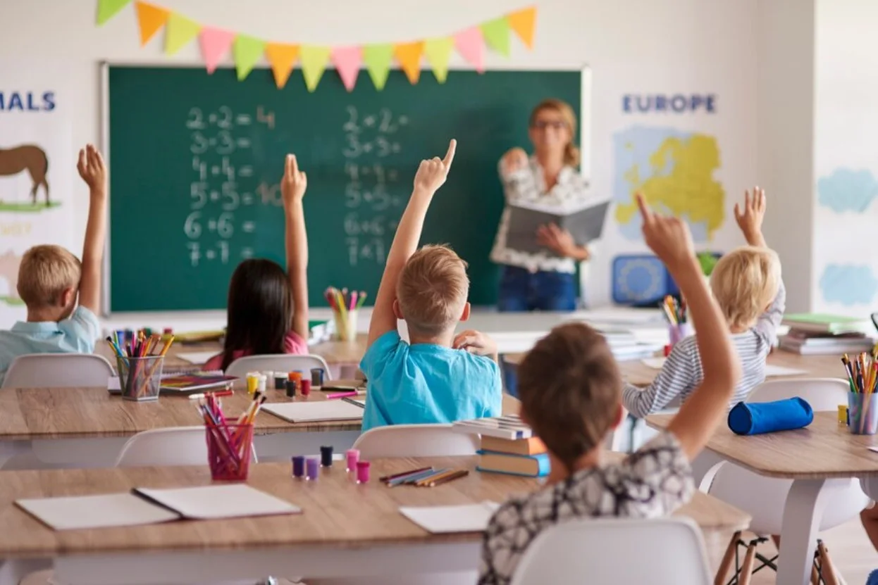 Young students raising hands in the classroom with the teacher by the chalkboard
